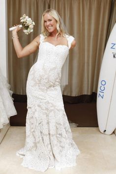 a woman in a white wedding dress standing next to a surfboard and holding flowers