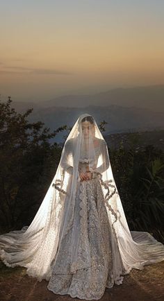 a woman wearing a wedding dress and veil standing on top of a hill at sunset