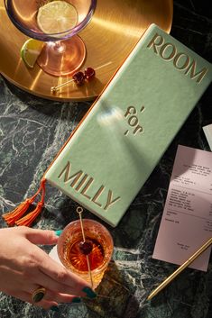 a person holding a cocktail in front of a room for milly book on a table