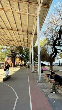 two people sitting on benches under a covered area next to a basketball court with trees in the background