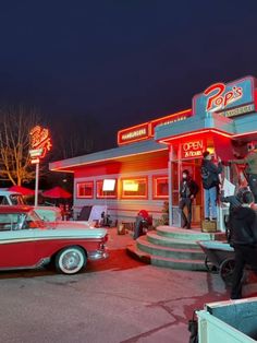 an old car is parked in front of a diner at night with people standing outside