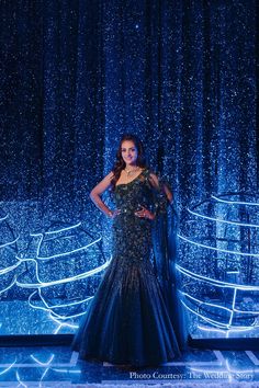 a woman standing in front of a blue backdrop wearing a black and gold dress with sequins