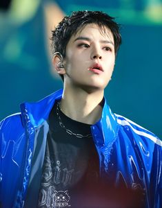 a young man with black hair wearing a blue jacket and ear piercings, looking off to the side