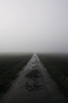 an empty road in the middle of a field on a foggy, overcast day