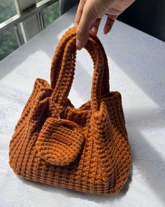 a hand holding an orange purse on top of a white tablecloth covered table next to a window