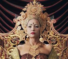 a woman in an elaborate dress sitting on a gold throne with red velvet drapes