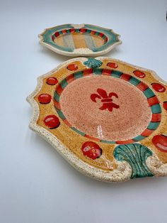 two decorative plates sitting on top of a white table next to each other, one with an orange and green design