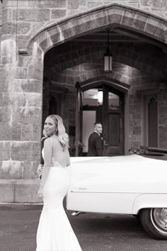 a woman standing next to a white car in front of a stone building with an arched doorway