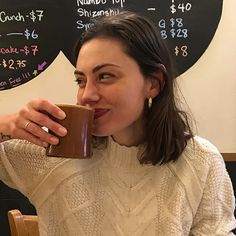 a woman sitting at a table drinking out of a coffee cup with chalk writing on the wall behind her