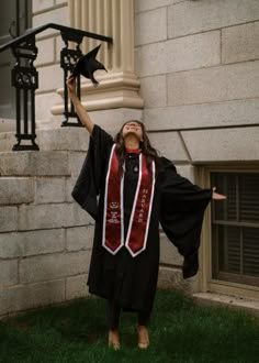 a woman wearing a graduation gown and holding her arms up in the air while standing on some grass