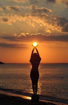 a woman is standing on the beach with her hands up in the air as the sun sets