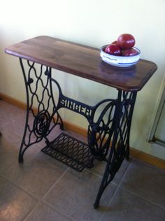 an old sewing table with two apples on top and a bowl of fruit sitting on it