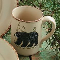 a coffee cup with a bear painted on it next to plates and pine branches in the background