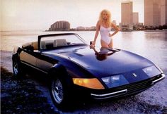 a woman standing on top of a black sports car next to the ocean with buildings in the background