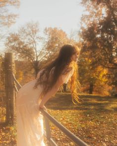 a woman in a white dress is leaning on a fence and looking at the ground