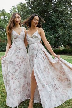 two women in long dresses standing next to each other on the grass with trees in the background