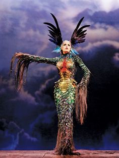 a woman dressed in an elaborate costume with feathers on her head and hands outstretched, standing against a cloudy sky