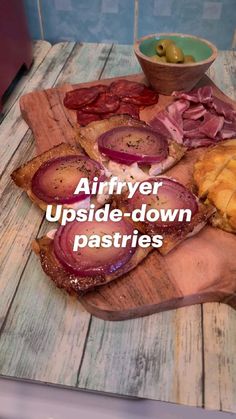 a wooden cutting board topped with sliced up meats and bread next to a bowl of olives