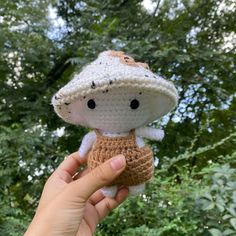 a hand holding a small crocheted doll in front of trees