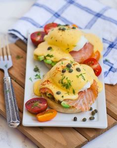 two eggs benedicts with tomatoes and capers on a white plate next to a fork