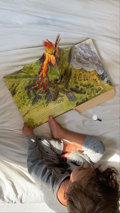 a little boy laying on top of a bed reading a book with an image of a volcano