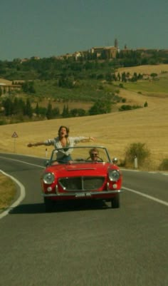 two people riding in the back of a red convertible car on a road with hills and fields behind them