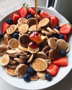 a white plate topped with pancakes and fruit next to a cup of syrup being poured on top