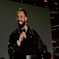 a man standing in front of a large screen holding a microphone and wearing a black jacket