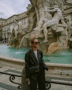 a woman standing in front of a fountain with statues on the sides and people walking around