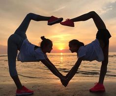 two women doing yoga poses on the beach at sunset with their hands in the shape of a heart