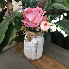 a mason jar filled with pink and white flowers on top of a wooden table next to potted plants