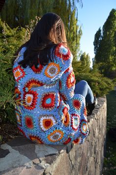 a woman sitting on a stone wall wearing a colorful crochet jacket and jeans