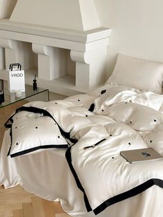 an unmade bed with white sheets and black piping on the comforter is in front of a fireplace