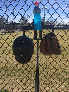 a baseball glove and helmet hanging on a chain link fence with a water bottle in the background