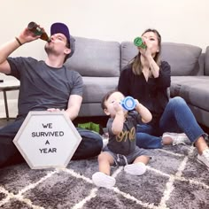 a man and woman sitting on the floor with a baby drinking from bottles next to a sign that says we survived a year