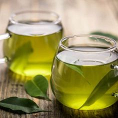 two mugs filled with green tea sitting on top of a wooden table next to leaves