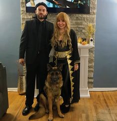 a man and woman standing next to a dog in front of a fireplace with a painting on the wall behind them