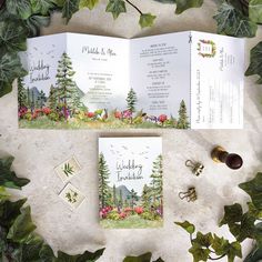 two wedding cards with trees and flowers on them, sitting next to some greenery