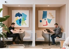 two people sitting at tables with laptops in front of paintings on the wall behind them