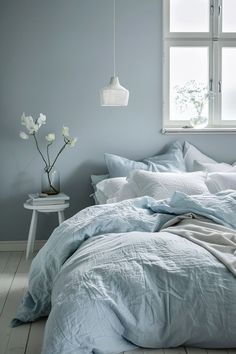 an unmade bed with blue sheets and white flowers in the corner next to a window