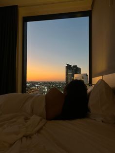 a person laying on a bed in front of a window looking out at the city