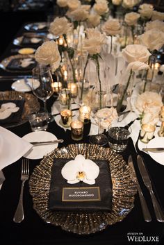 a black table topped with white flowers and silver place settings, candles and napkins