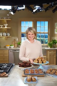 a woman standing in a kitchen holding a plate of food next to trays of cookies