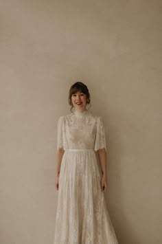 a woman standing in front of a wall wearing a white dress and smiling at the camera