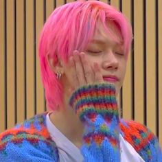 a person with pink hair wearing a colorful sweater and mittens holding their hands to their face