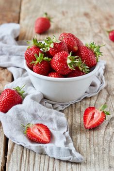 a white bowl filled with strawberries sitting on top of a wooden table next to a cloth