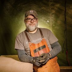 a man with an apron and hat is standing in front of a green wall holding a piece of wood