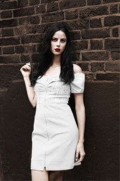 a woman with long black hair wearing a white dress standing in front of a brick wall