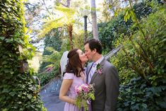 a bride and groom kissing in front of greenery at their wedding ceremony, with the bride wearing a pink dress