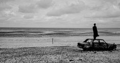 a man standing on top of an old car in the sand at the beach near the ocean